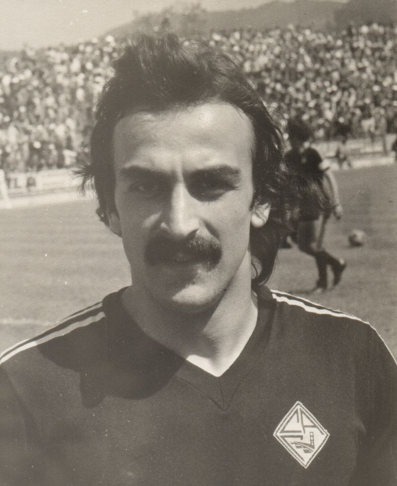 Black and white portrait of a young José Costa, with long hair and a moustache, in the black Académica shirt, with a crowd behind him