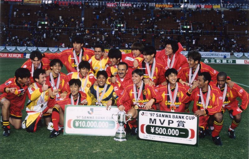 Nagoya Grampus Eight players and staff celebrating on the pitch with the 1997 Sanwa Bank Cup trophy and prize boards