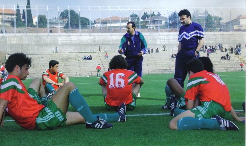 Two coaches standing on a training pitch while players in Portugal kit sit on the grass
