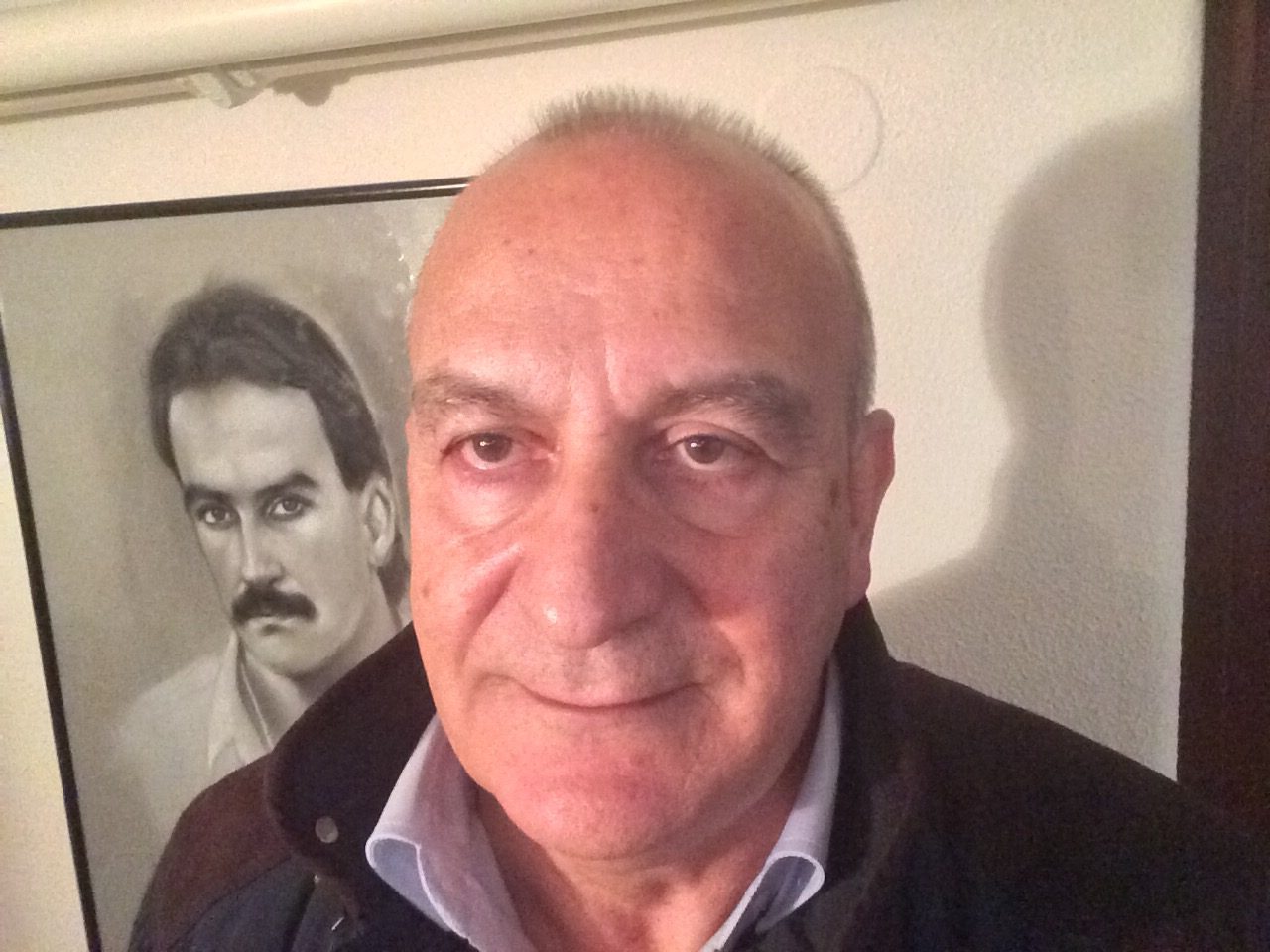 José Costa today, smiling, in front of a framed drawing of a player with long hair and a moustache