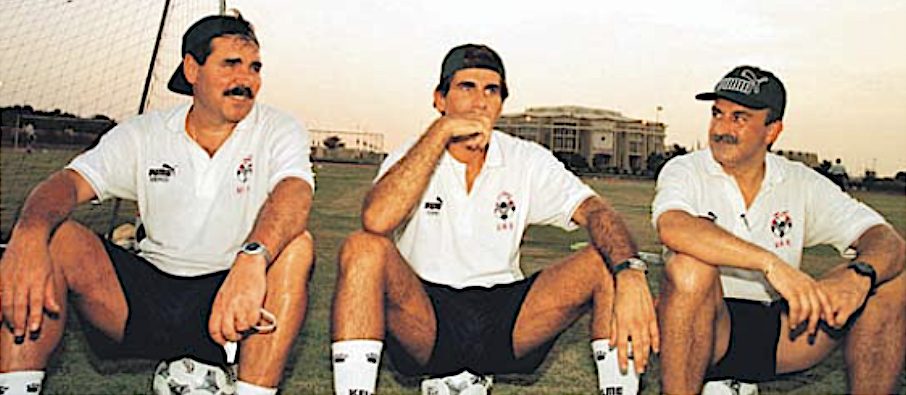 Three coaches in white training kit and caps sitting on a training pitch in Abu Dhabi